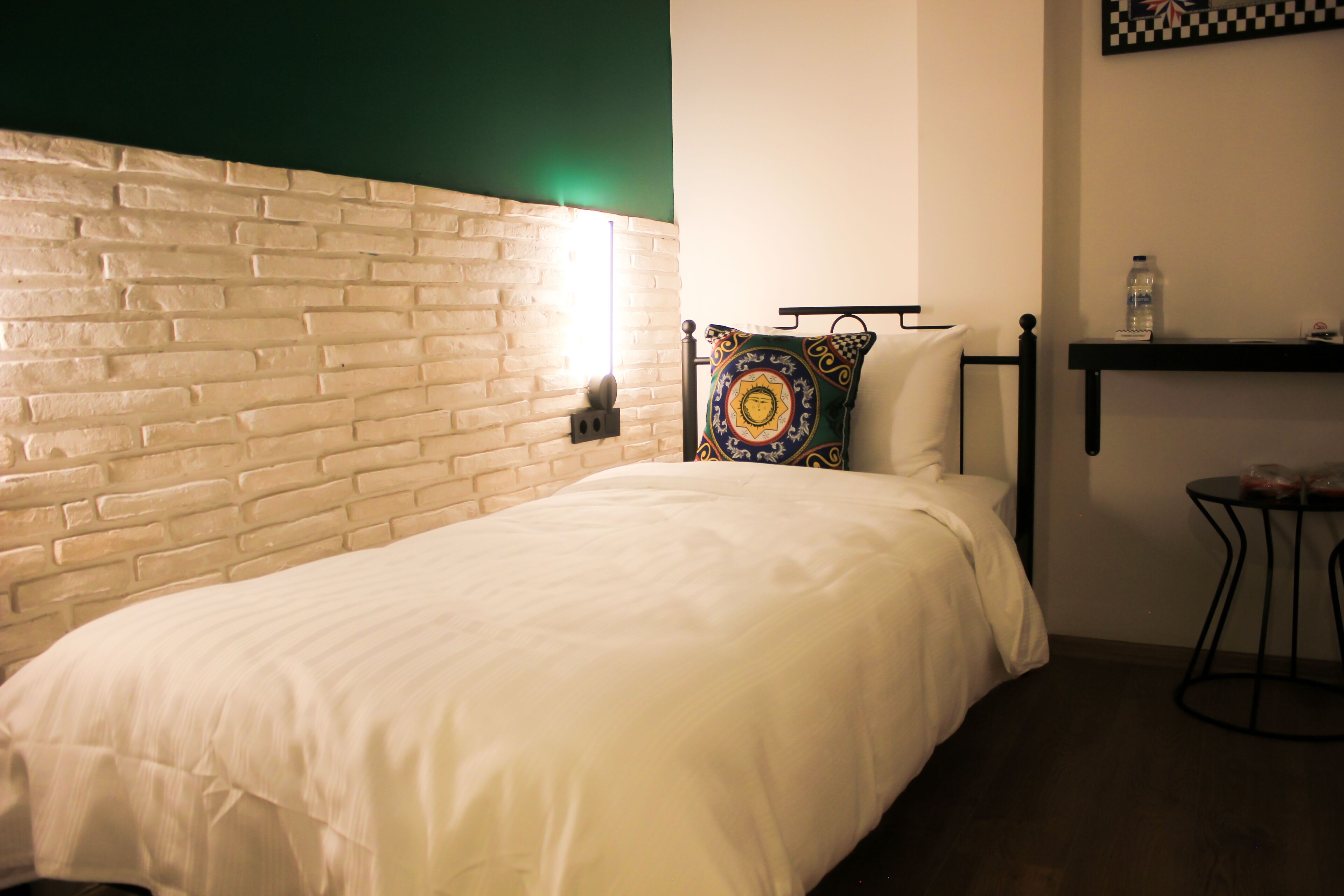 A room with a white brick headboard, a white bedspread, a colorful pillow, a small black table with a water bottle, and a shelf with a checkered pattern.