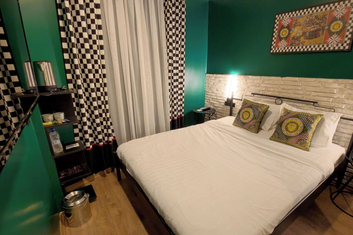 A hotel room with a checkered curtain, a bed with patterned pillows, a small table, and a wall-mounted painting.