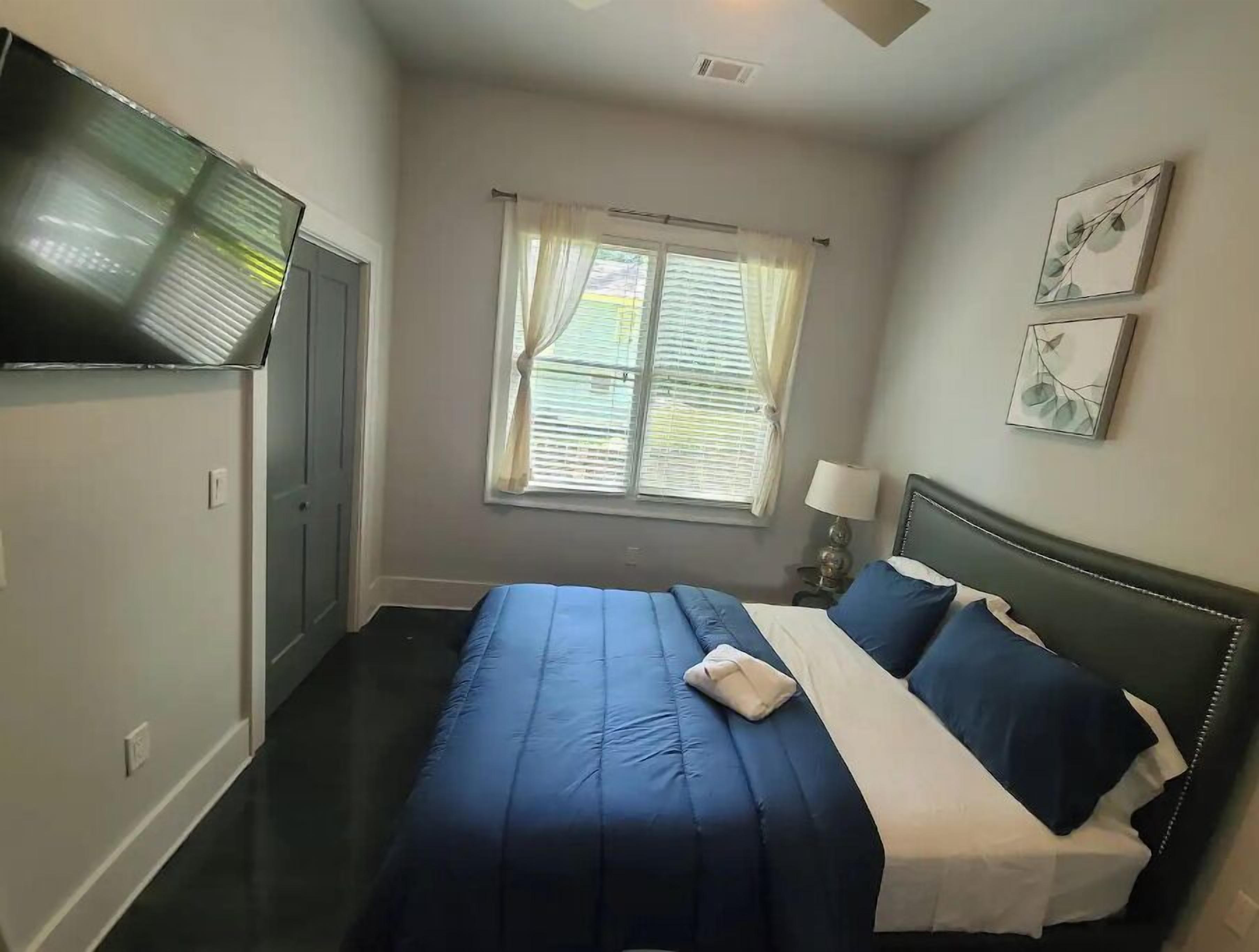 A hotel room with a bed, a window with curtains, a flat-screen TV mounted on the wall, and a small table with a lamp.