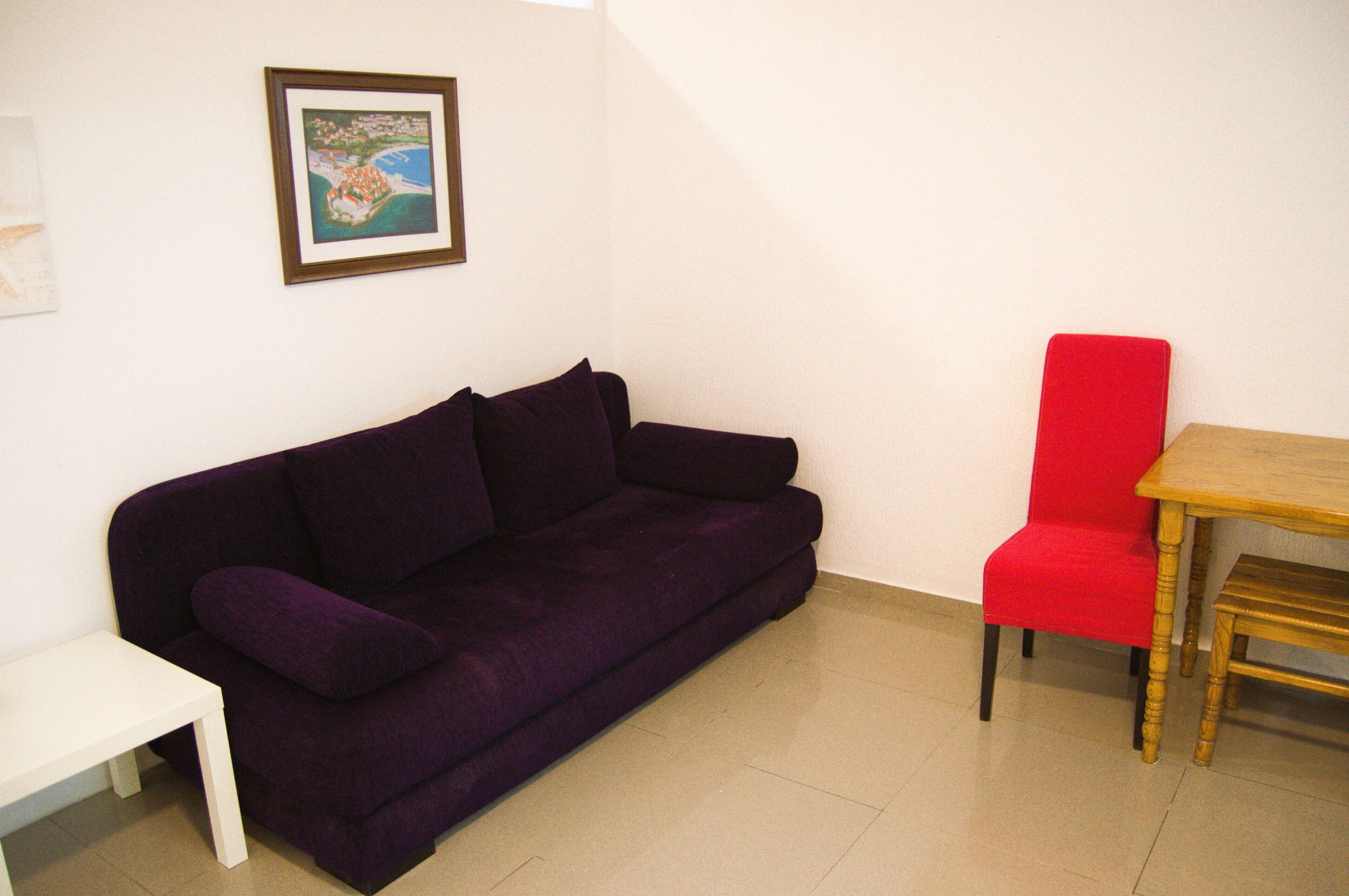 Ein violettes Sofa, ein roter Sessel und ein kleiner weißer Tisch in einem Raum mit weißen Wänden.