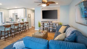 Ein modernes Wohnzimmer mit einem blauen Sofa, einem Couchtisch und einem Fernseher. Es gibt eine Küchenzeile mit weißen Schränken und einem Esstisch mit Stühlen.