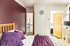 A bedroom with a wooden bed, purple bedding, a clock on the wall, and a TV on the right.
