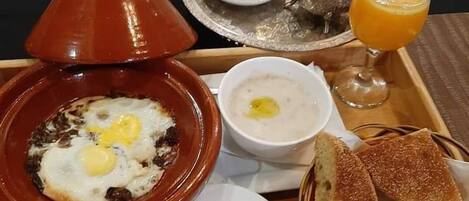 A breakfast tray with a tagine, soup, bread, and a glass of orange juice.