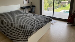 Quarto com uma cama grande, uma escrivaninha e um violão. Vê-se um jardim e montanhas pela janela.