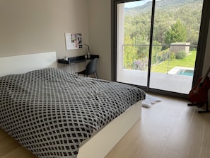 Une chambre avec un grand lit, un bureau et une guitare. La fenêtre offre une vue sur un jardin et des montagnes.