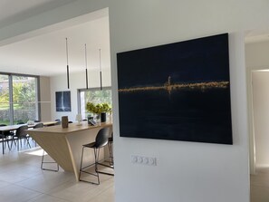 A modern kitchen with a large island, pendant lights, and a cityscape painting.