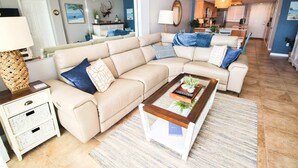 Un salon avec un canapé d’angle beige, une table basse et une table d’appoint avec une lampe. Au fond, on aperçoit une cuisine avec un mur bleu et un luminaire suspendu.