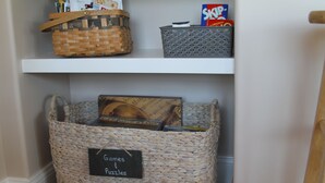 Un espace de rangement avec des paniers en osier étiquetés « Jeux et casse-têtes » et une étagère au-dessus contenant une boîte de céréales Skips et un autre panier.