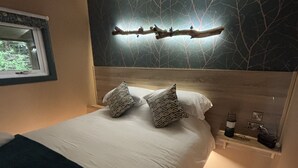 A bedroom with a bed, pillows, a nightstand, a wall-mounted light fixture, and a window. - Heywood Glamping (Ormskirk)
