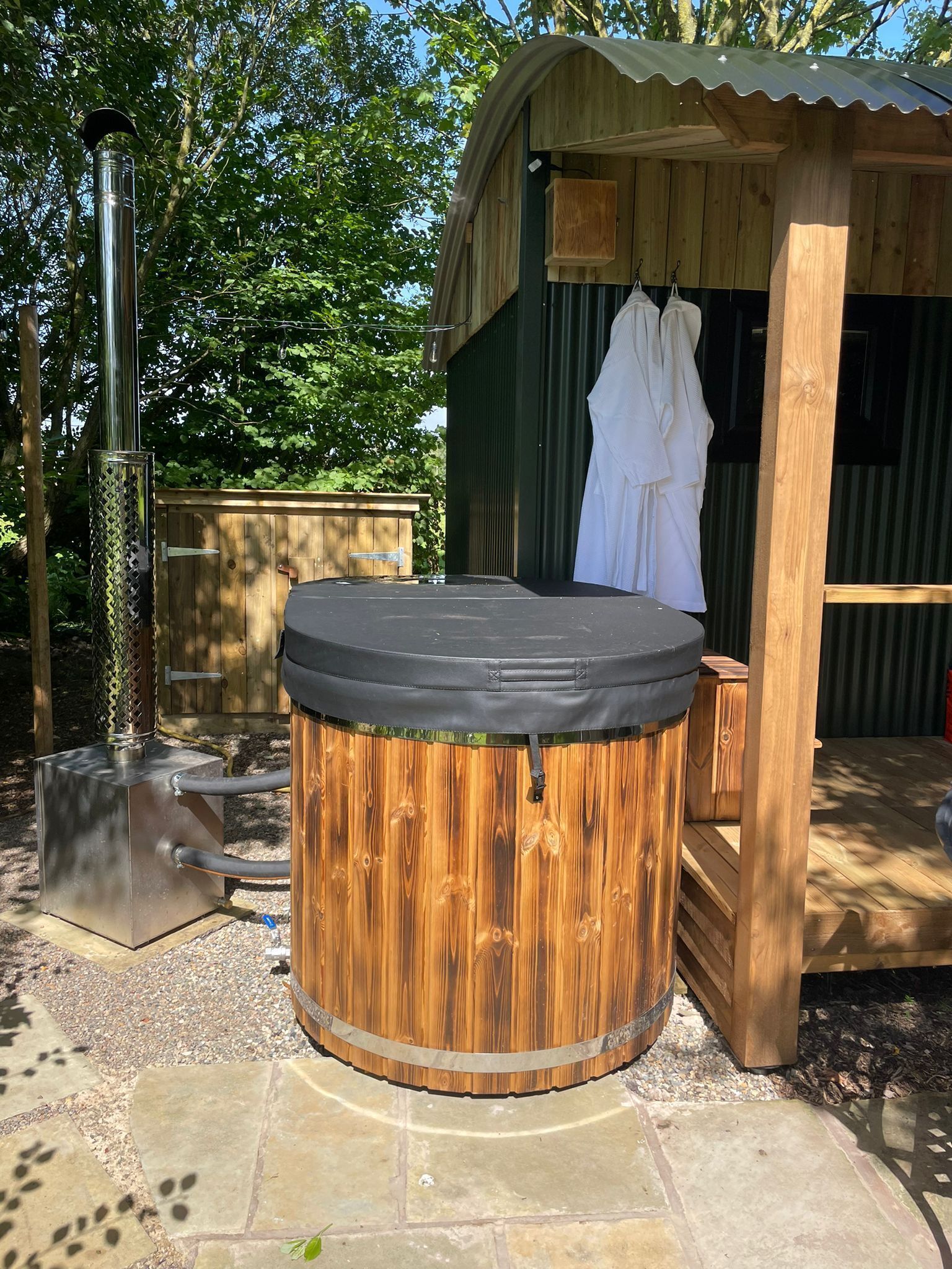 A wooden hot tub with a metal heater, a wooden deck, and a covered wooden structure with a white towel hanging.