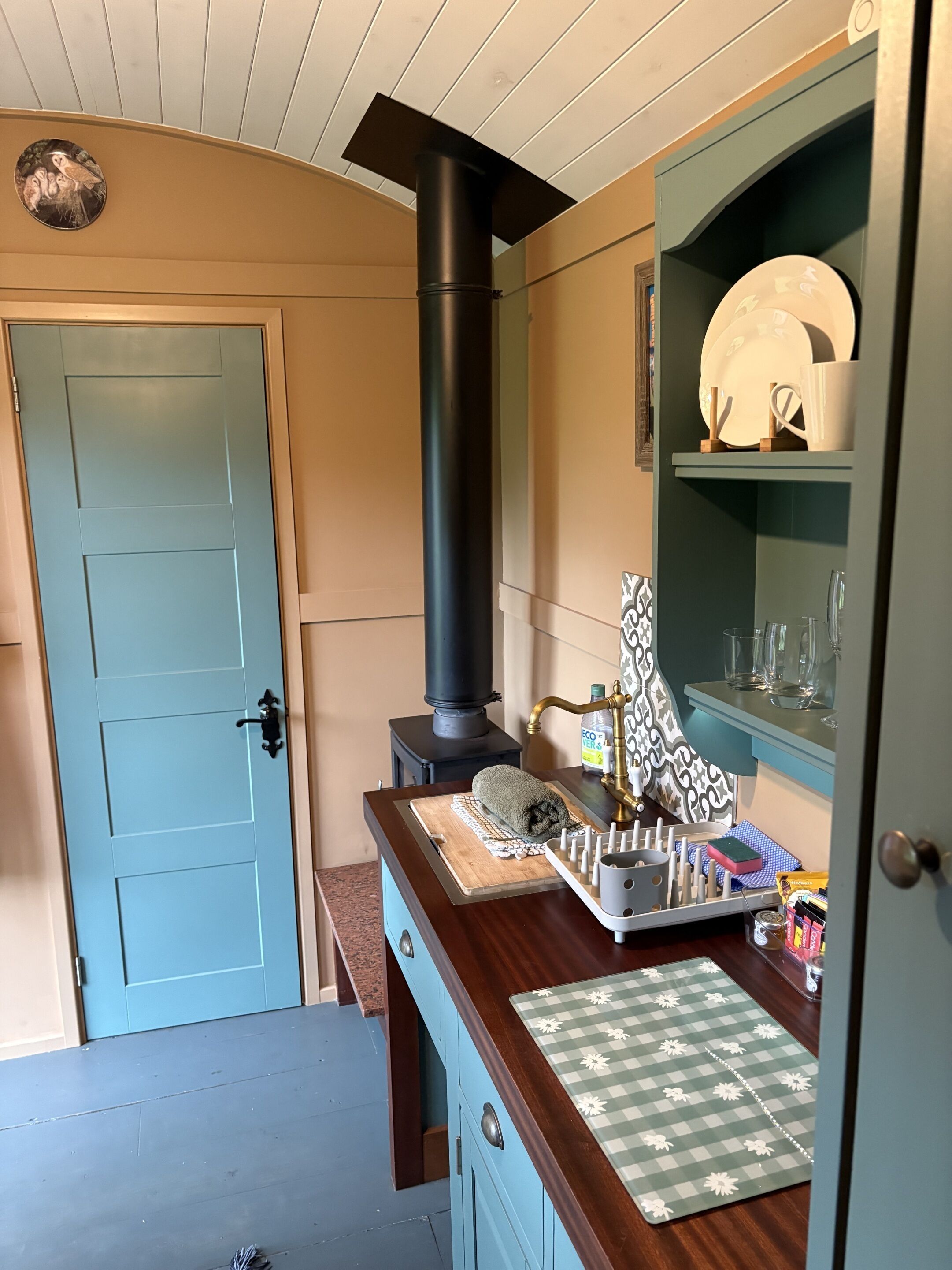Een kamer met een blauwe deur, een zwarte schoorsteen, een wastafel met een decoratieve achterwand en een plank met servies en glazen.