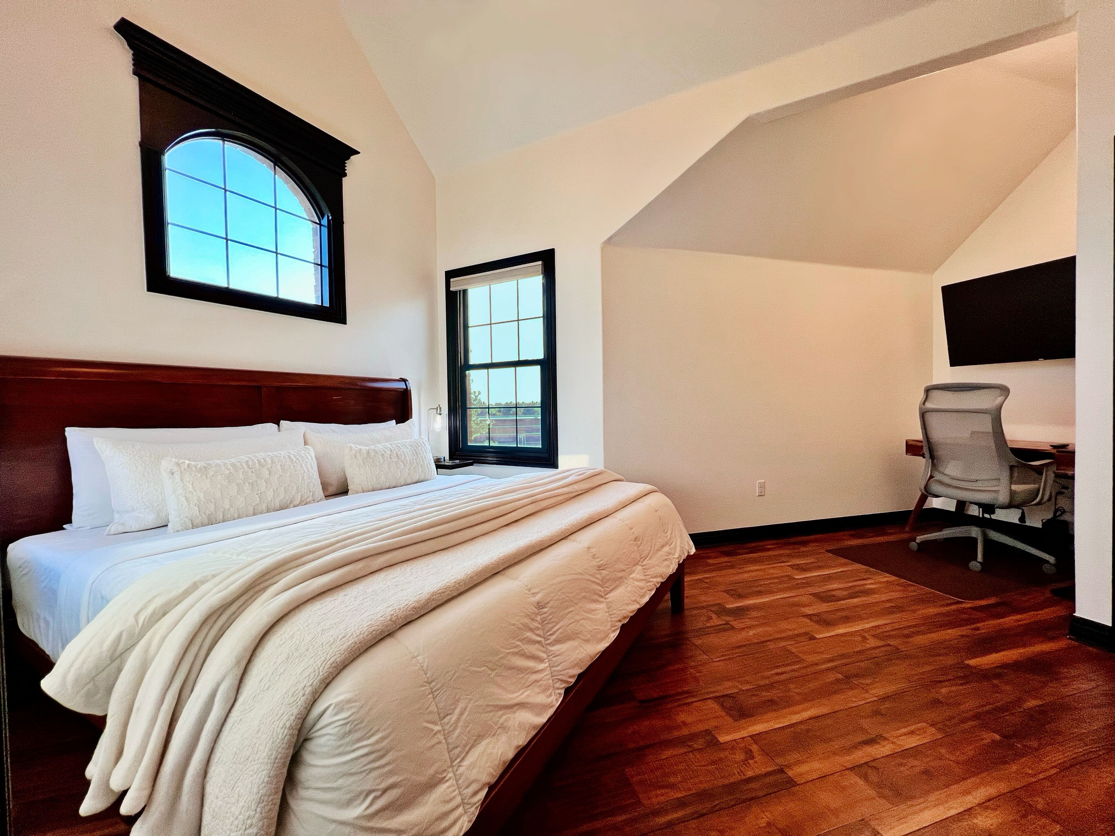 A bedroom with a large bed, a desk with a chair, and two windows.