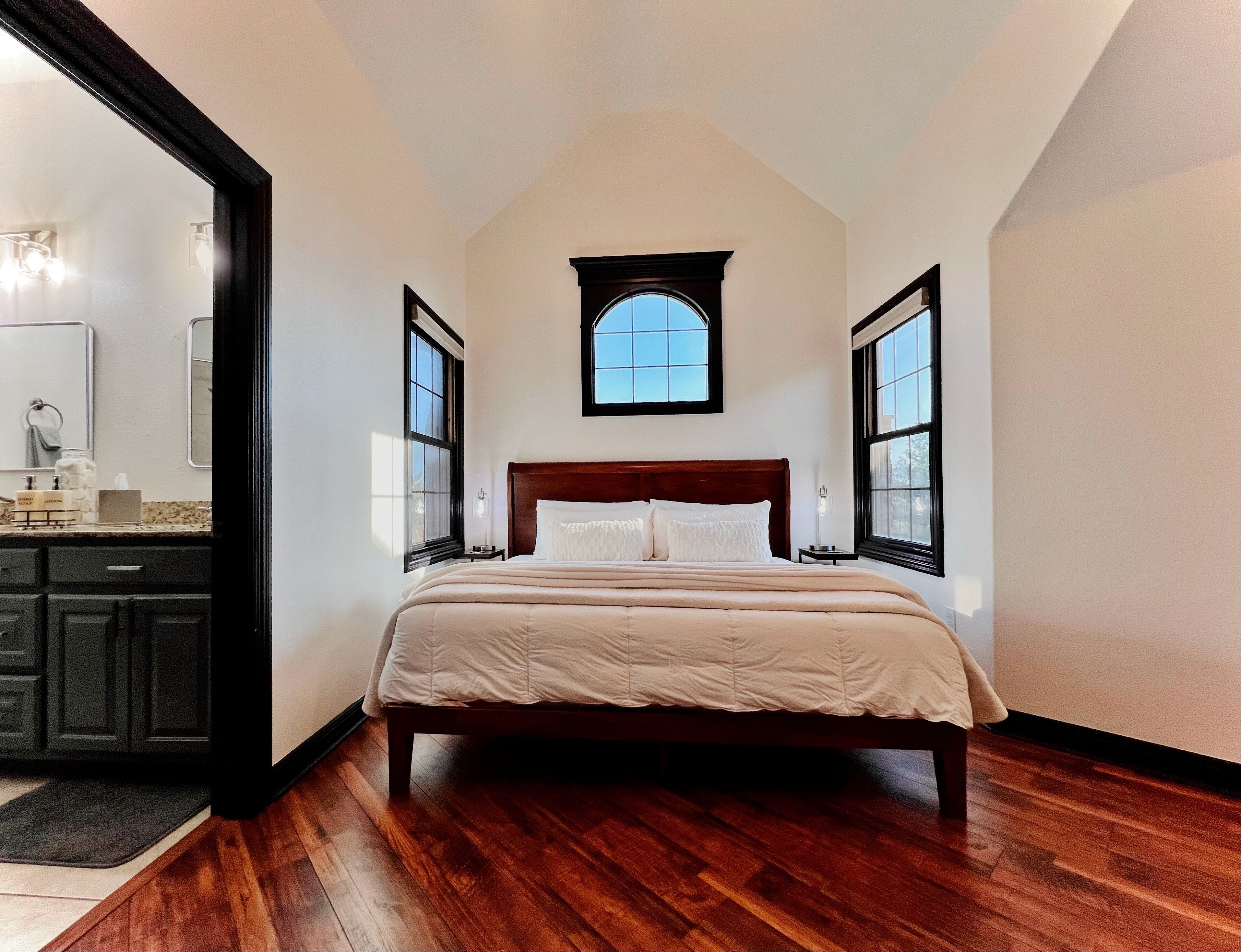 Un dormitorio con una cama de madera, un ventanal pequeño y un baño visible a través de una puerta abierta.