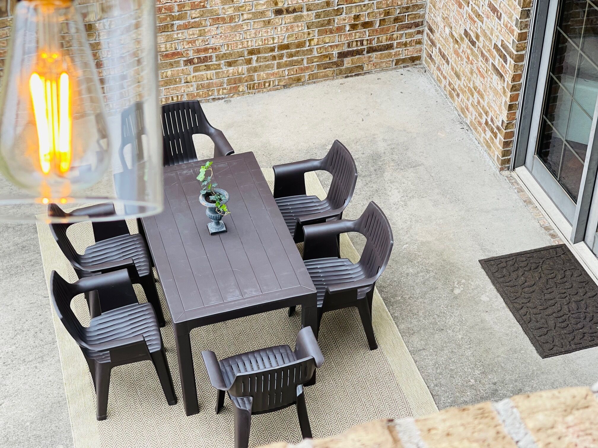An outdoor patio with a dark wooden table and black plastic chairs, a potted plant, and a brick wall in the background.