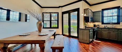 A modern kitchen with a wooden dining table, a vase with branches, and a view through the large sliding glass door.