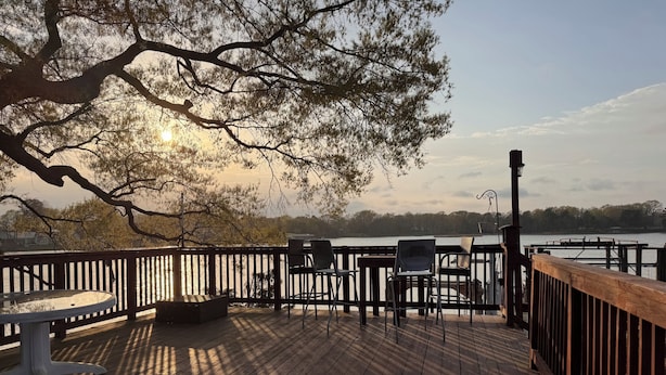 Une terrasse en bois, avec des chaises et une table, donnant sur une étendue d’eau, avec des arbres en arrière-plan.