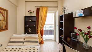 Habitación de hotel con cama, escritorio con ordenador, estantería y ventana con cortinas.