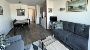 Una sala moderna con un sofá gris, una mesa de comedor con sillas, una zona de cocina con televisor y un cuadro en la pared.