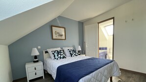 Un dormitorio con techo inclinado, una cama con manta azul, dos mesitas de noche con lámparas, una puerta que da a un balcón y un cuadro enmarcado en la pared.