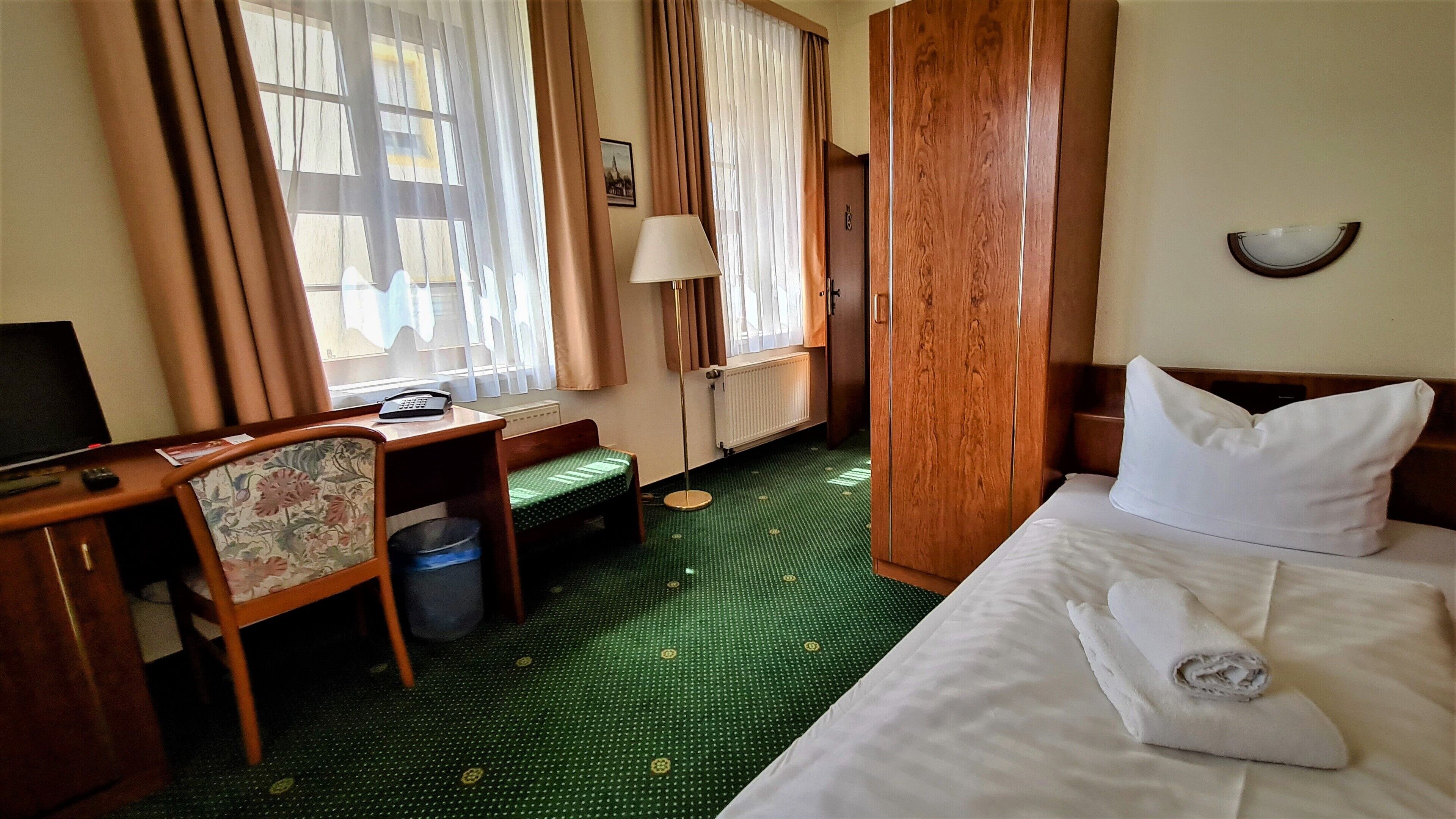 Habitación de hotel con cama, escritorio, silla, lámpara, radiador y ventana con cortinas.