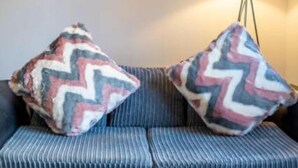 Ein Zimmer mit einer gestreiften Couch, zwei gemusterten Kissen, einer Stehlampe und einer Vase mit Blumen auf einem Glastisch.