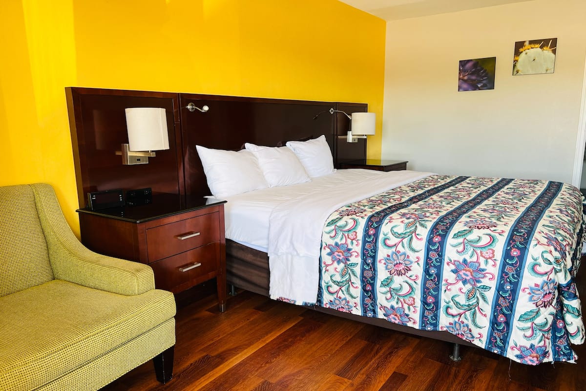 Une chambre d’hôtel avec une tête de lit en bois, un lit recouvert d’une courtepointe à motifs floraux, une table de chevet avec une lampe et une chaise.