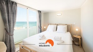 Un cuarto de hotel con cama, una mesita de noche con lámpara, vista a la playa y balcón con cortinas.