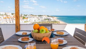 Una mesa servida con alimentos para el desayuno, incluyendo un bol de fruta y pasteles, con vistas a un pueblo costero y una playa.