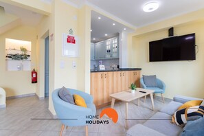 Een moderne woonkamer met een bank, fauteuils, een salontafel en een televisie. Er is een keukenhoek met kasten en een spoelbak. Een brandblusser is aan de muur bevestigd.