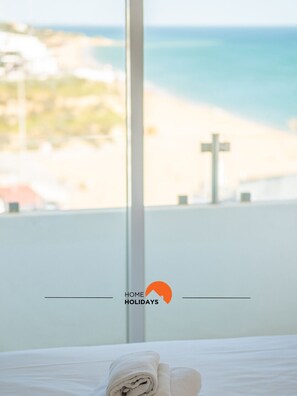 Hotelkamer met groot raam met uitzicht op een strand en een kruis, met een bed en een opgerolde handdoek.