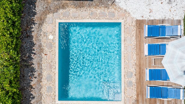 Vista aérea de una piscina rectangular con borde de piedra, rodeada por un sendero de guijarros y sillas de jardín azules.