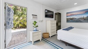 Un dormitorio con cama, televisión, cuadro, mesita de noche y vista a un jardín a través de una puerta.