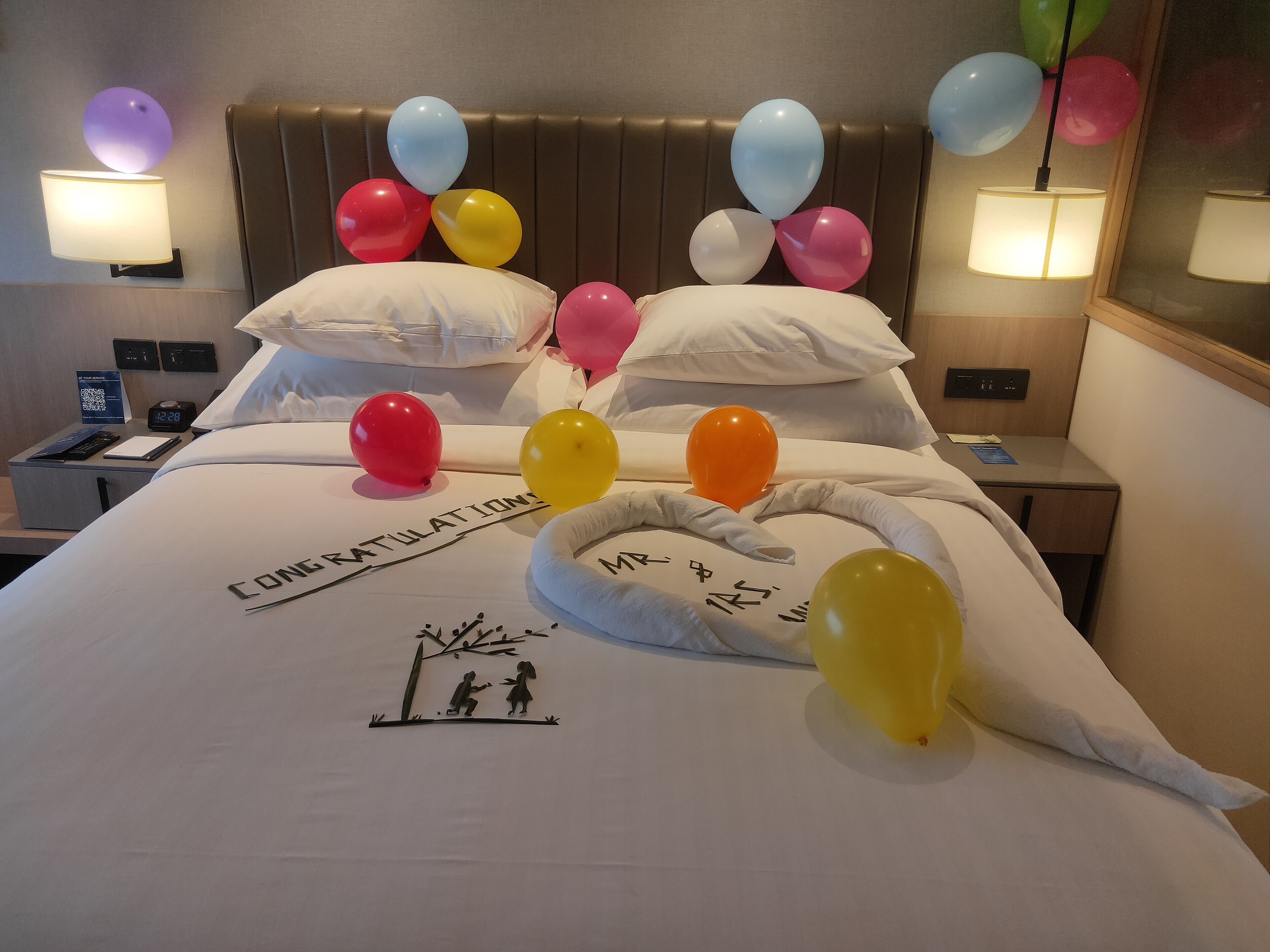 A hotel room with a bed, pillows, a bedside table, a nightstand, a lamp, a clock, and balloons.