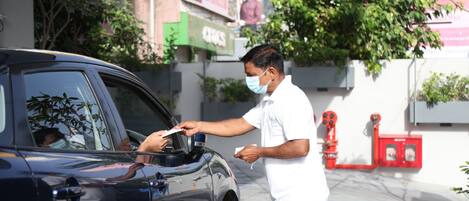 A man interacting with a car, possibly a service or inspection, in an urban setting with buildings and a tree in the background.