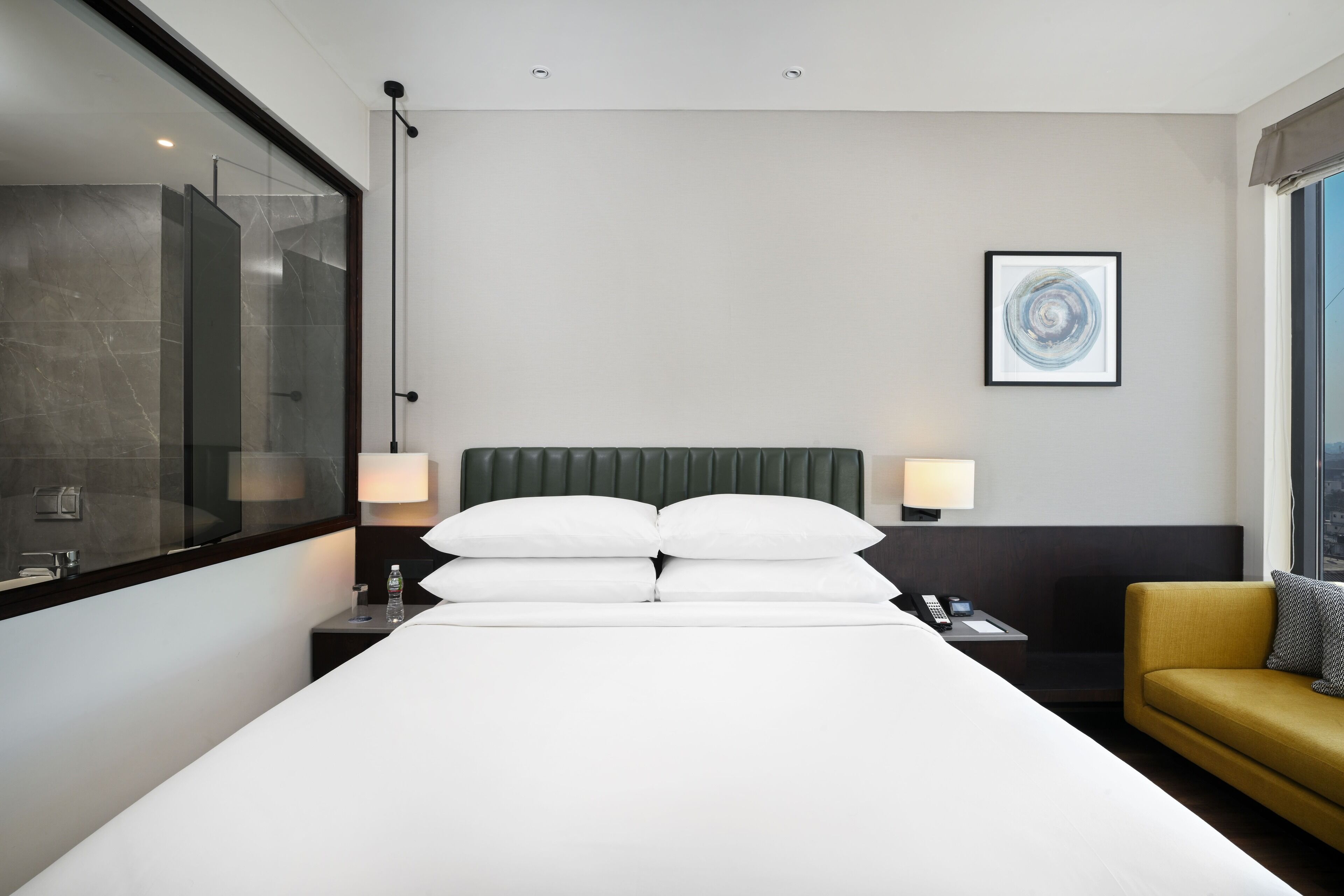 A hotel room with a large bed, a bedside table with a lamp, a small table with a phone, a sofa, and a window with a view.