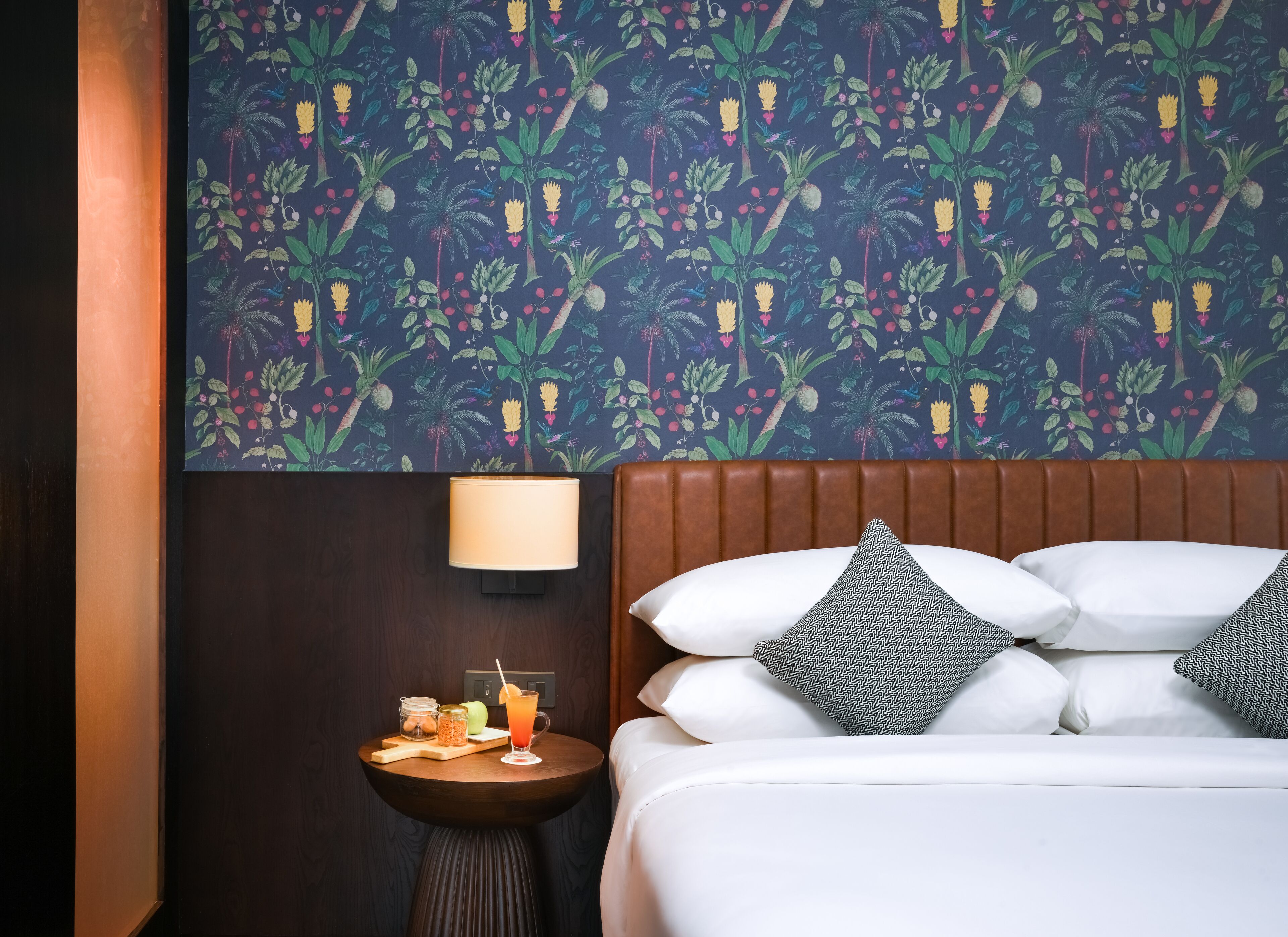 A hotel room with a bed, a nightstand, a lamp, and a wall with a tropical floral pattern.