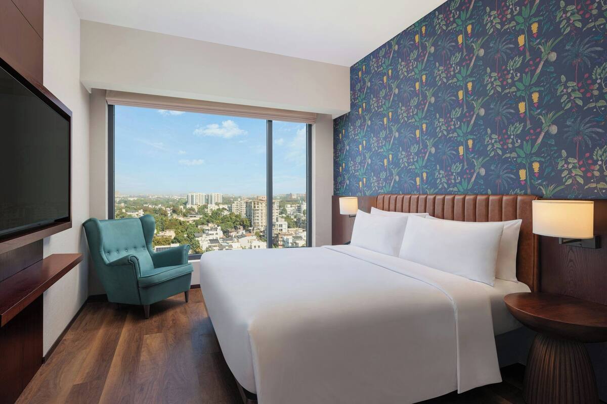 Une chambre d’hôtel avec un grand lit, un fauteuil turquoise, une télévision et une vue sur la ville.