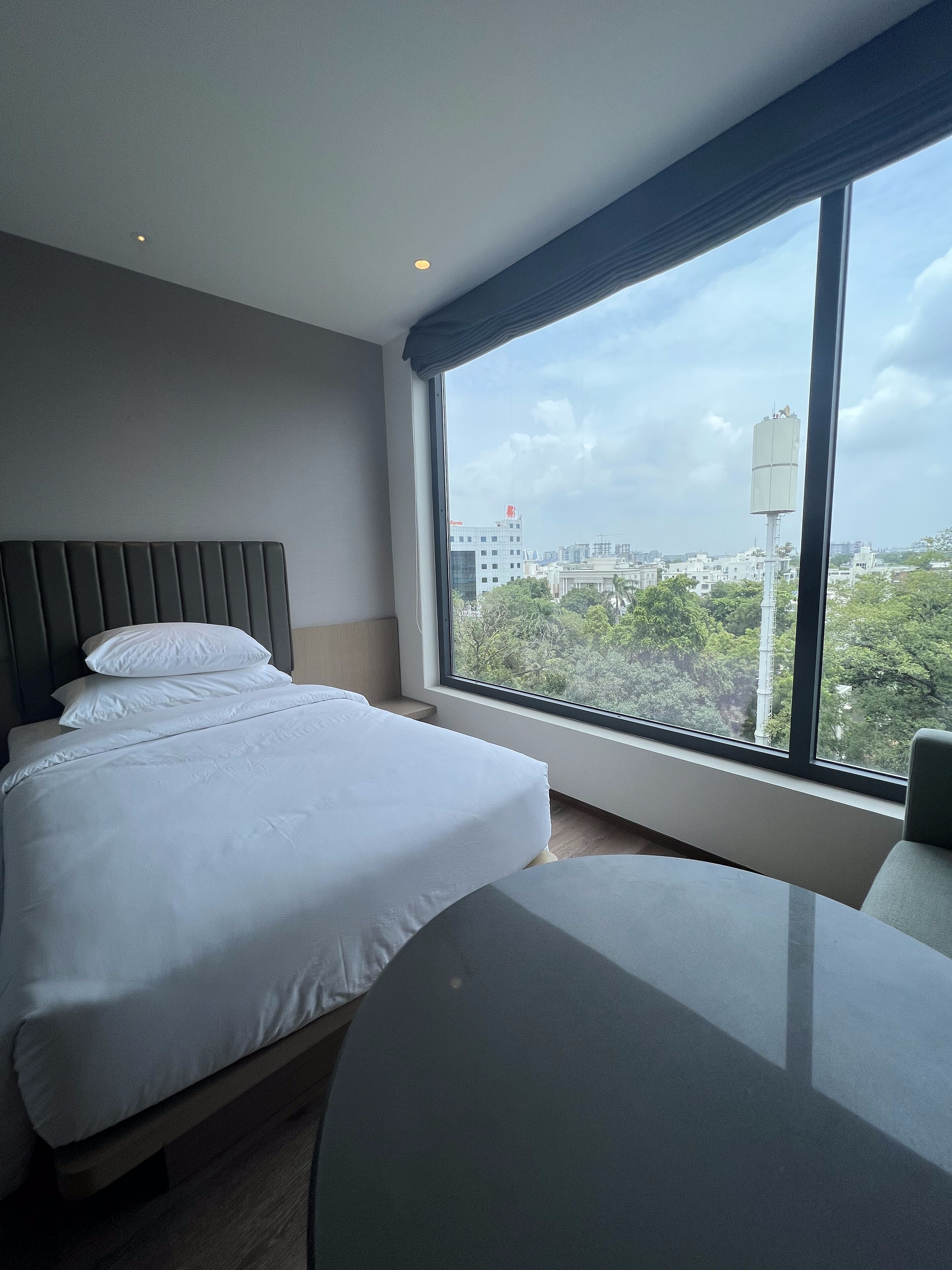 A modern hotel room with a large bed, a round table, and a view of the cityscape through a large window.