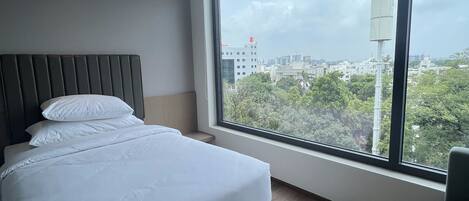 A modern hotel room with a large bed, a round table, and a view of the cityscape through a large window.