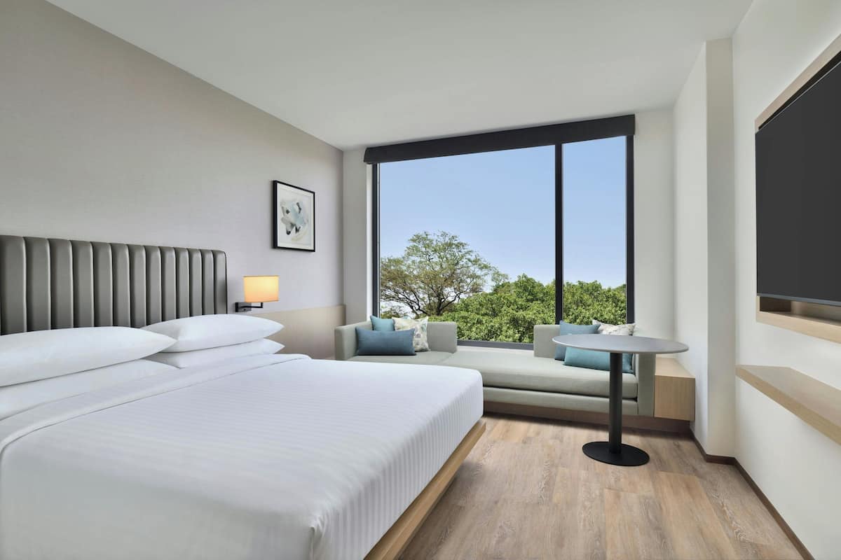 Une chambre d’hôtel moderne dotée d’un grand lit, d’un coin salon avec une table et d’une grande fenêtre donnant sur la verdure.
