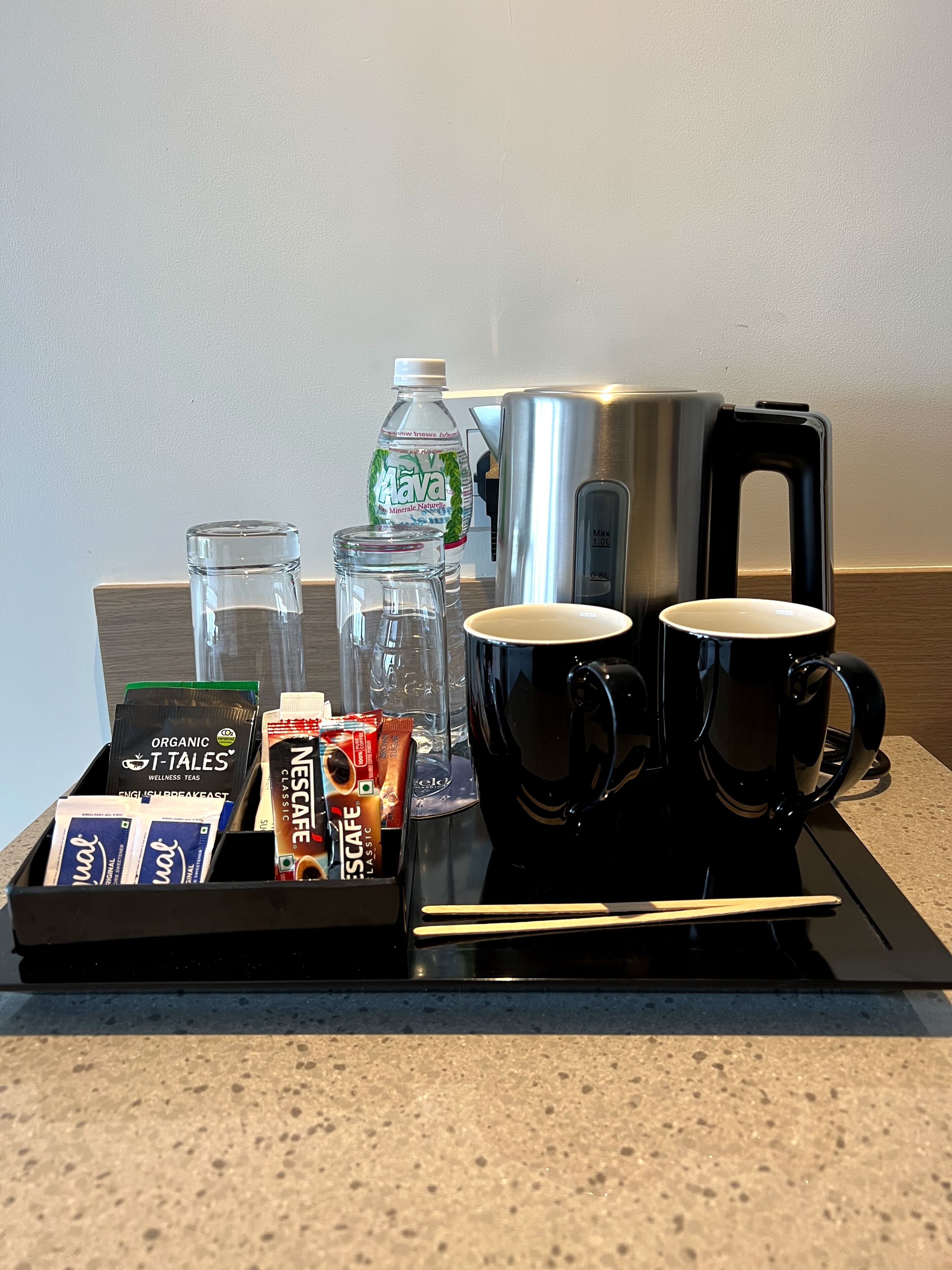 A coffee station with a coffee maker, two mugs, a water bottle, and a tray with tea packets and glasses.
