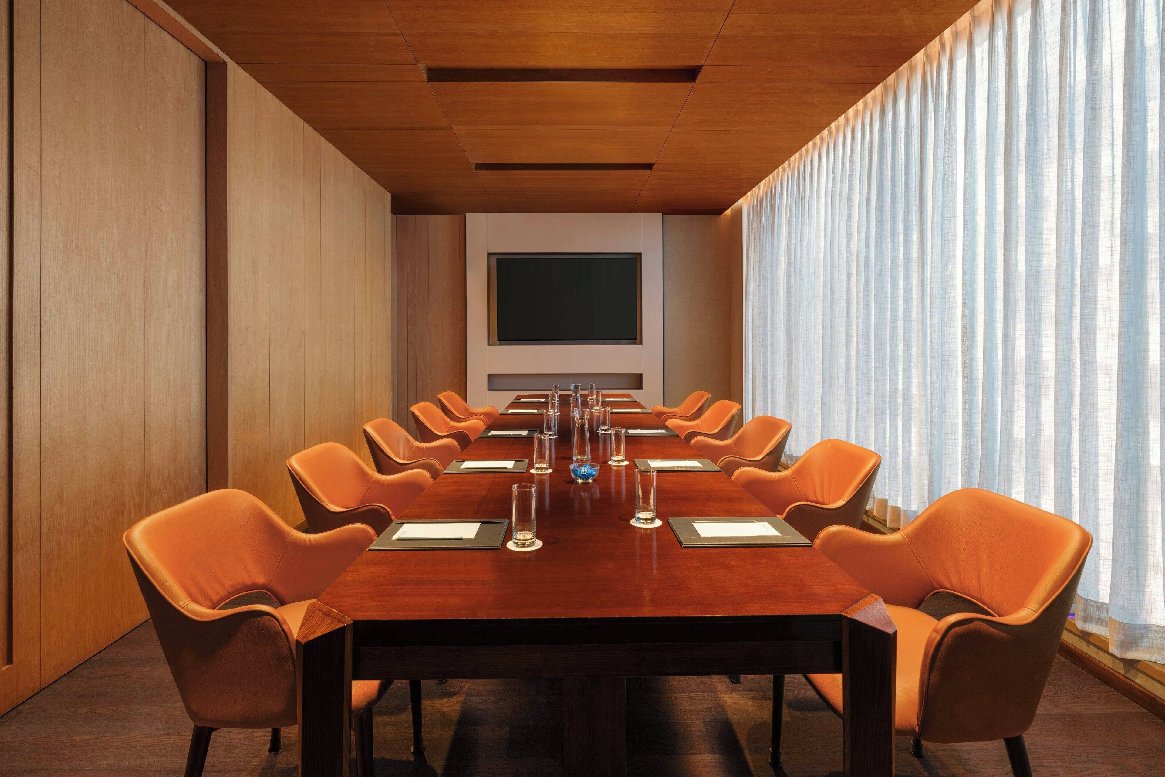 Een vergaderruimte met een lange tafel, oranje stoelen, een groot scherm en een raam met gordijnen.