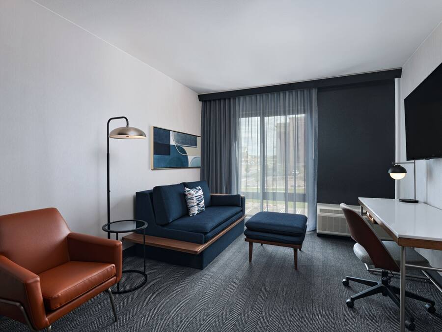 A modern hotel room with a sofa, armchair, desk, and large window.