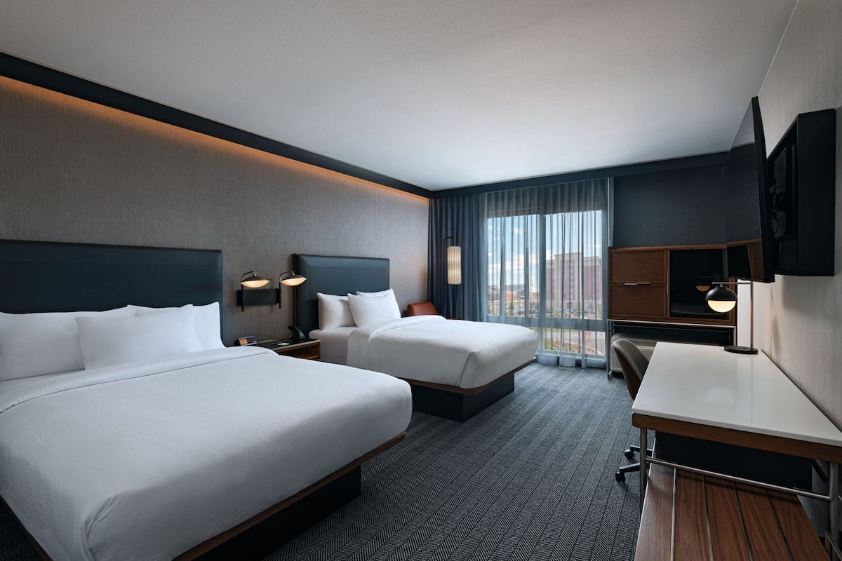 A hotel room with two beds, a desk, a chair, a TV, and a large window with curtains.