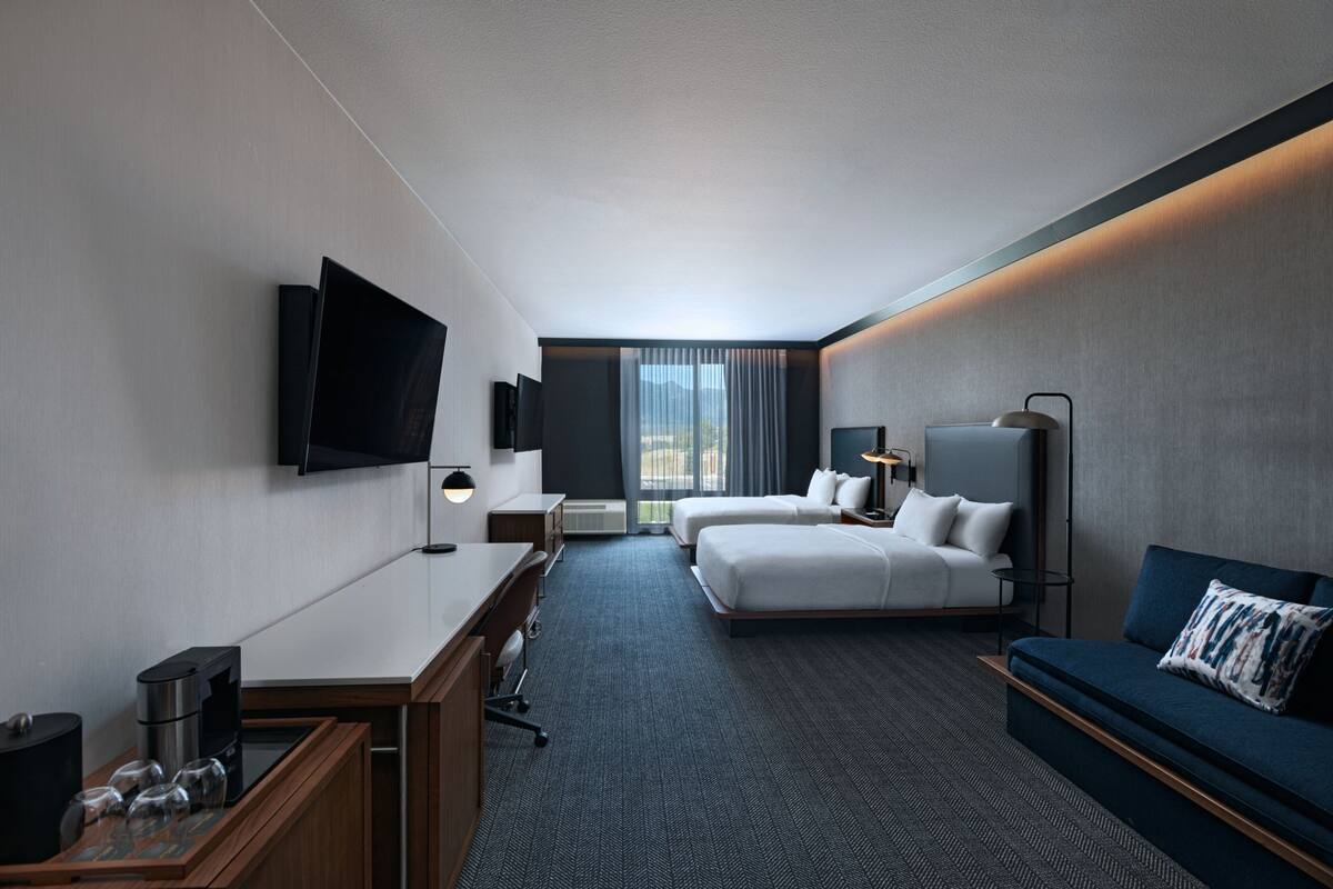 Una habitación de hotel moderna con televisión de pantalla plana, escritorio con cafetera, dos camas, sofá y ventana grande con cortinas.