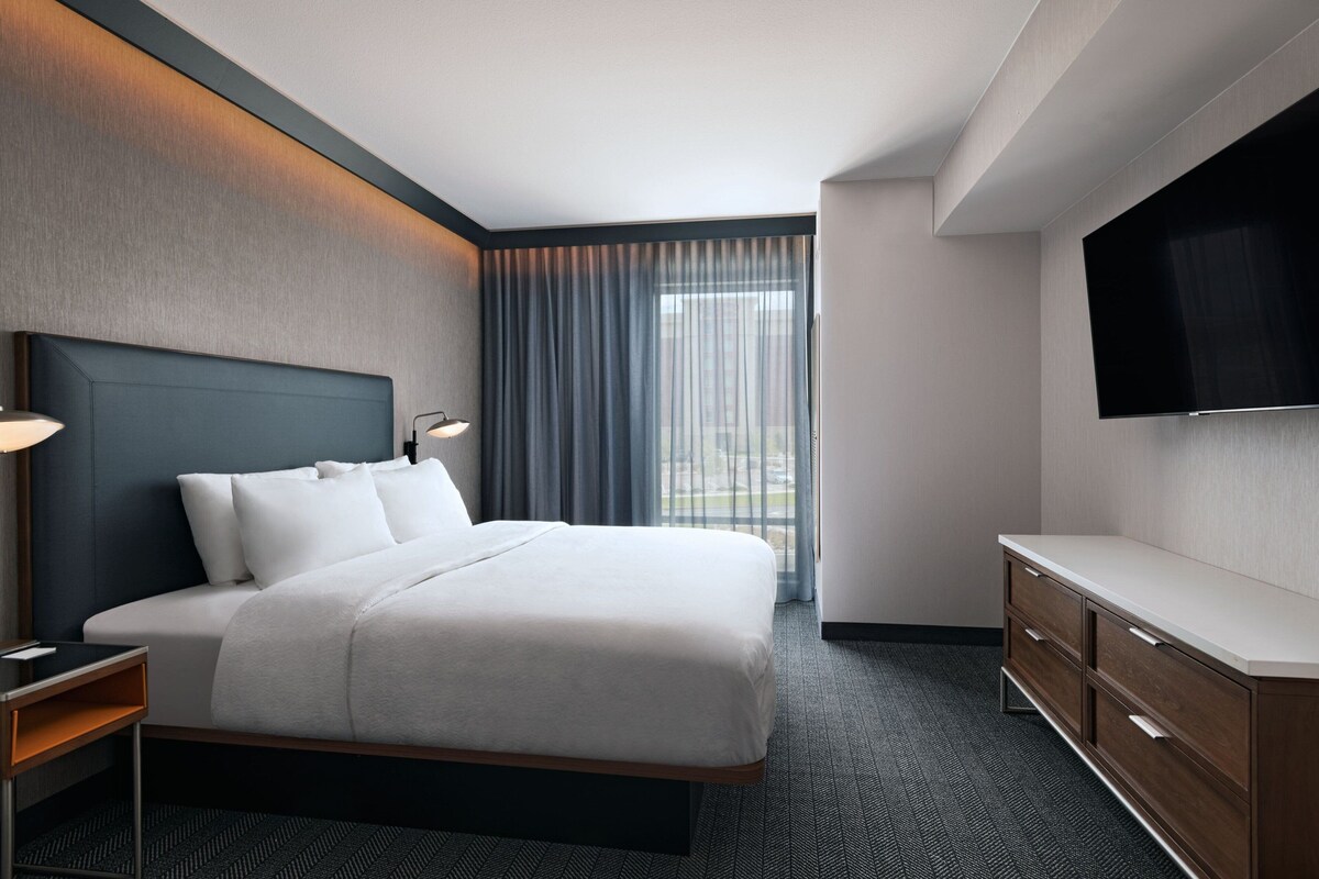 A modern hotel room with a large bed, a flat-screen TV, a wooden dresser, and a view of the city through the window.