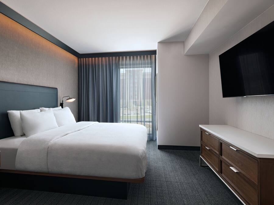 A modern hotel room with a large bed, a flat-screen TV, a wooden dresser, and a view of the city through the window.