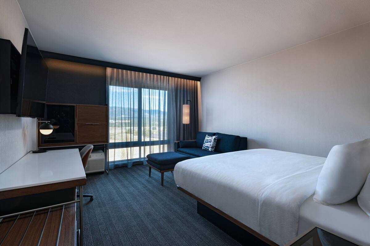 A hotel room with a bed, a desk, a chair, a TV, and a large window with curtains.