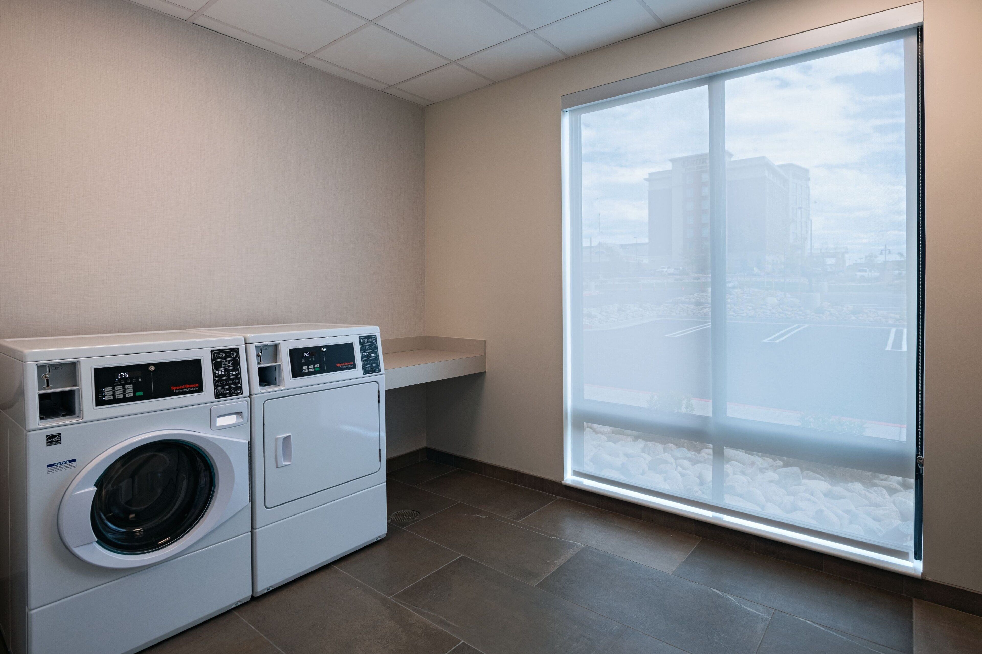 A laundry area with two front-loading washing machines, a small countertop, and a large window with a city view.