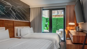 A hotel room with two beds, a TV, a desk, and a view of a green garden. - Le Meridien New York, Fifth Avenue (New York)